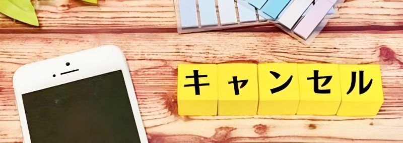 木目の机の上に置かれたスマートフォンと、「キャンセル」と書かれた黄色いブロックが並んでいる様子。
